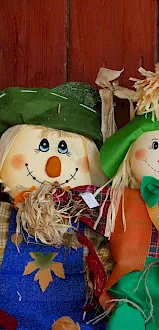 Two smiling scarecrow dolls with straw hair and green hats sit beside a large metal churn against a red wooden wall.